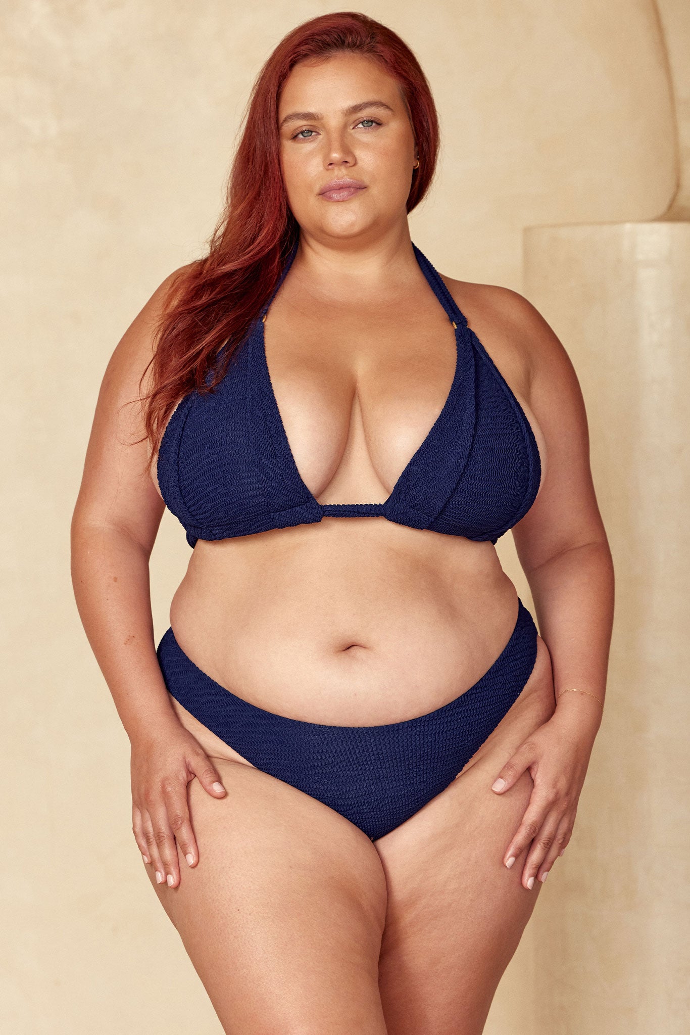 Plus-size woman with red hair standing in a navy bikini against a neutral backdrop, facing the camera.
