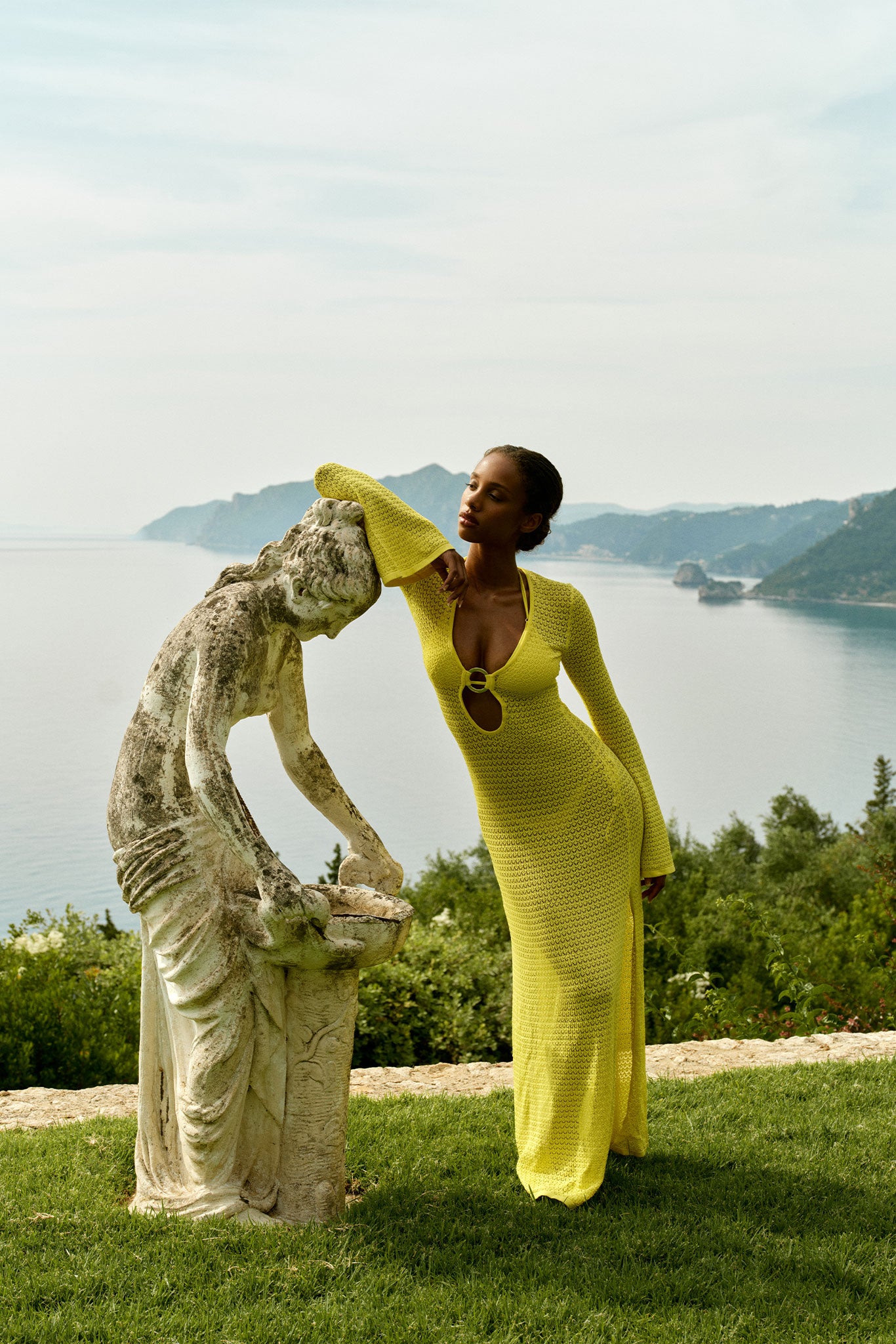 Woman in long yellow knit dress leaning on a weathered stone statue overlooking a coastal bay
