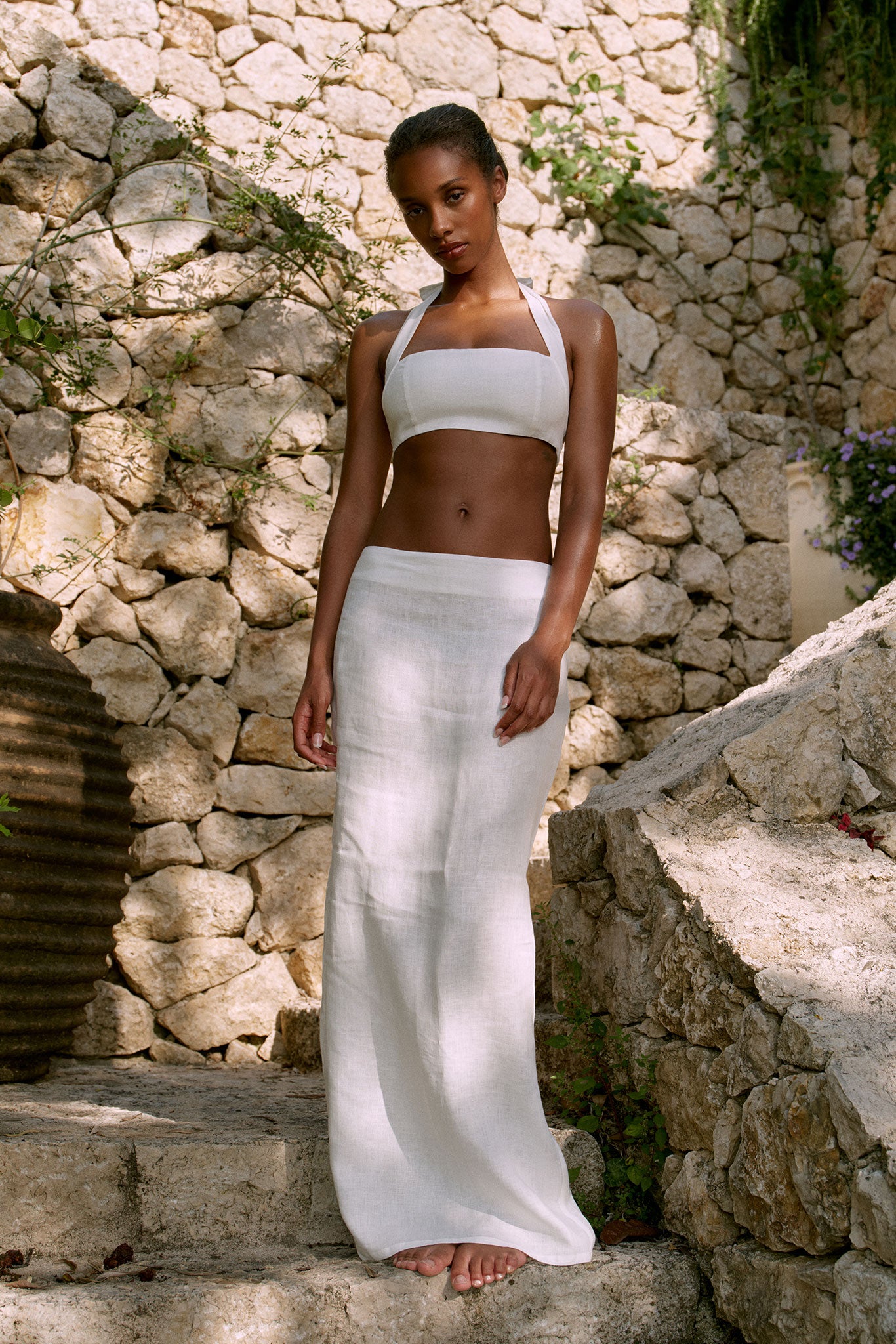 Woman wearing white linen halter crop top and long skirt, standing barefoot on stone steps against a sunlit rock wall