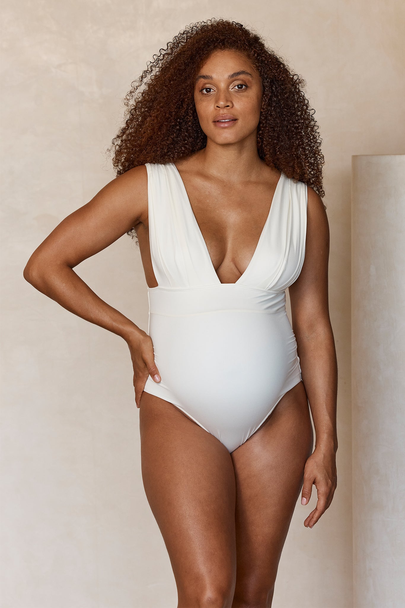 Pregnant woman wearing a plunging white one-piece swimsuit, standing against a beige background with hand on hip.