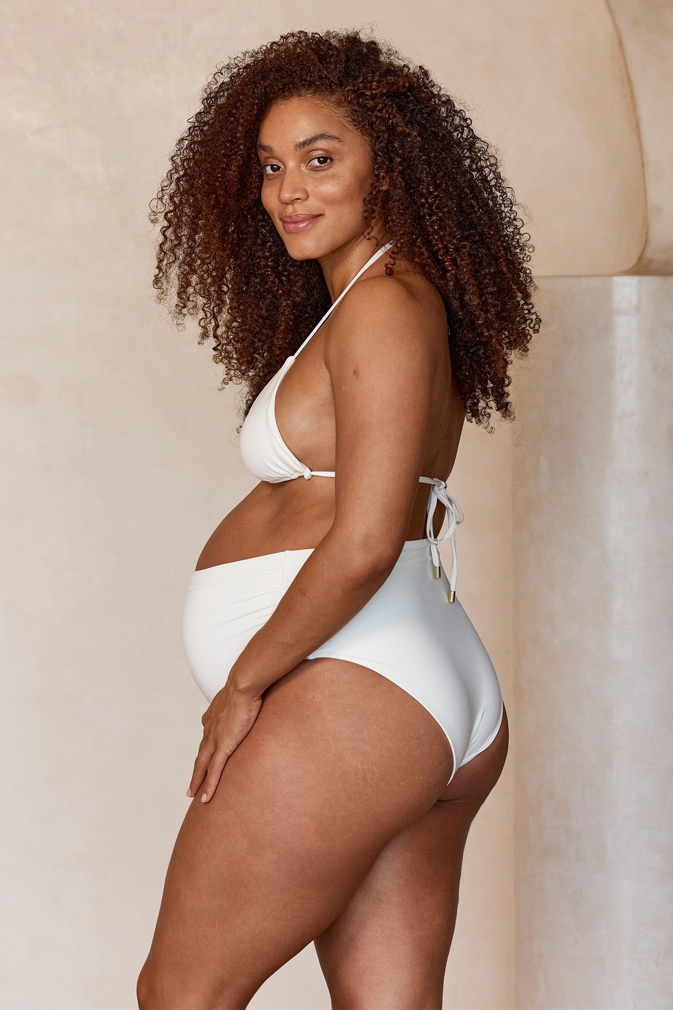 Pregnant woman modeling white two-piece maternity swimsuit, side profile, smiling, curly hair, hand resting on hip.