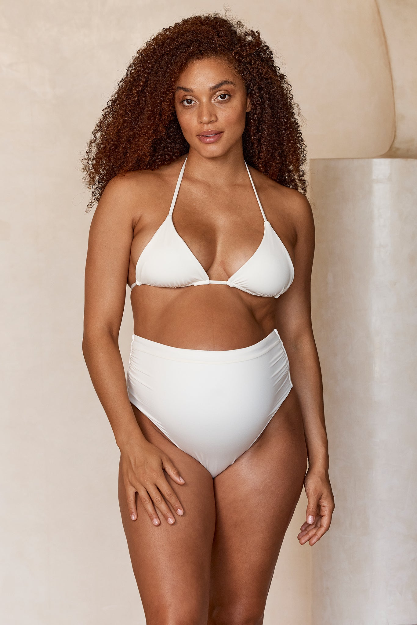Pregnant woman with curly hair wearing a white halter bikini and high‑waist bottoms, standing against a beige backdrop.