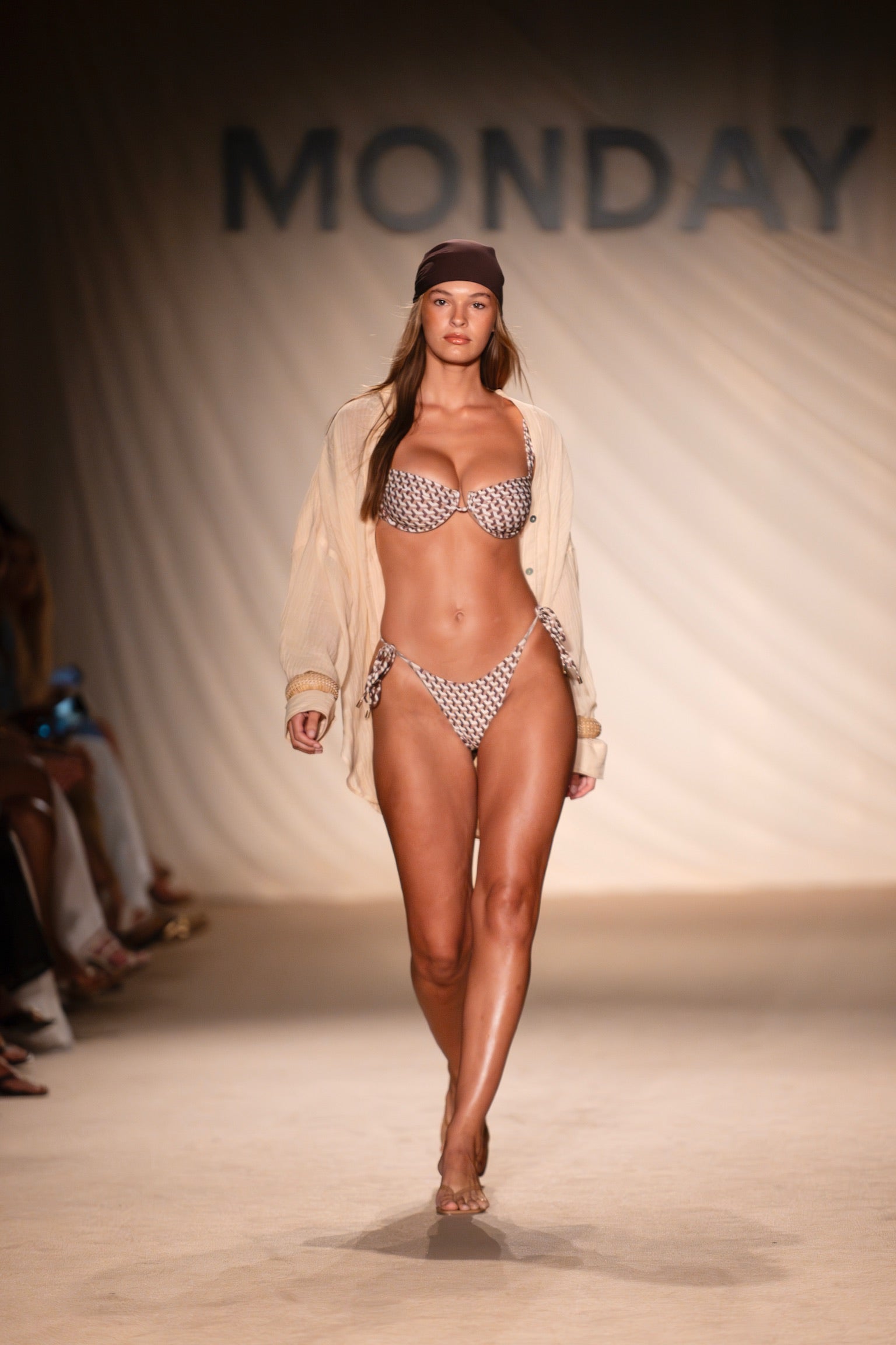 Runway model in patterned bikini, beige open shirt and brown headscarf walking down catwalk with 'MONDAY' backdrop.