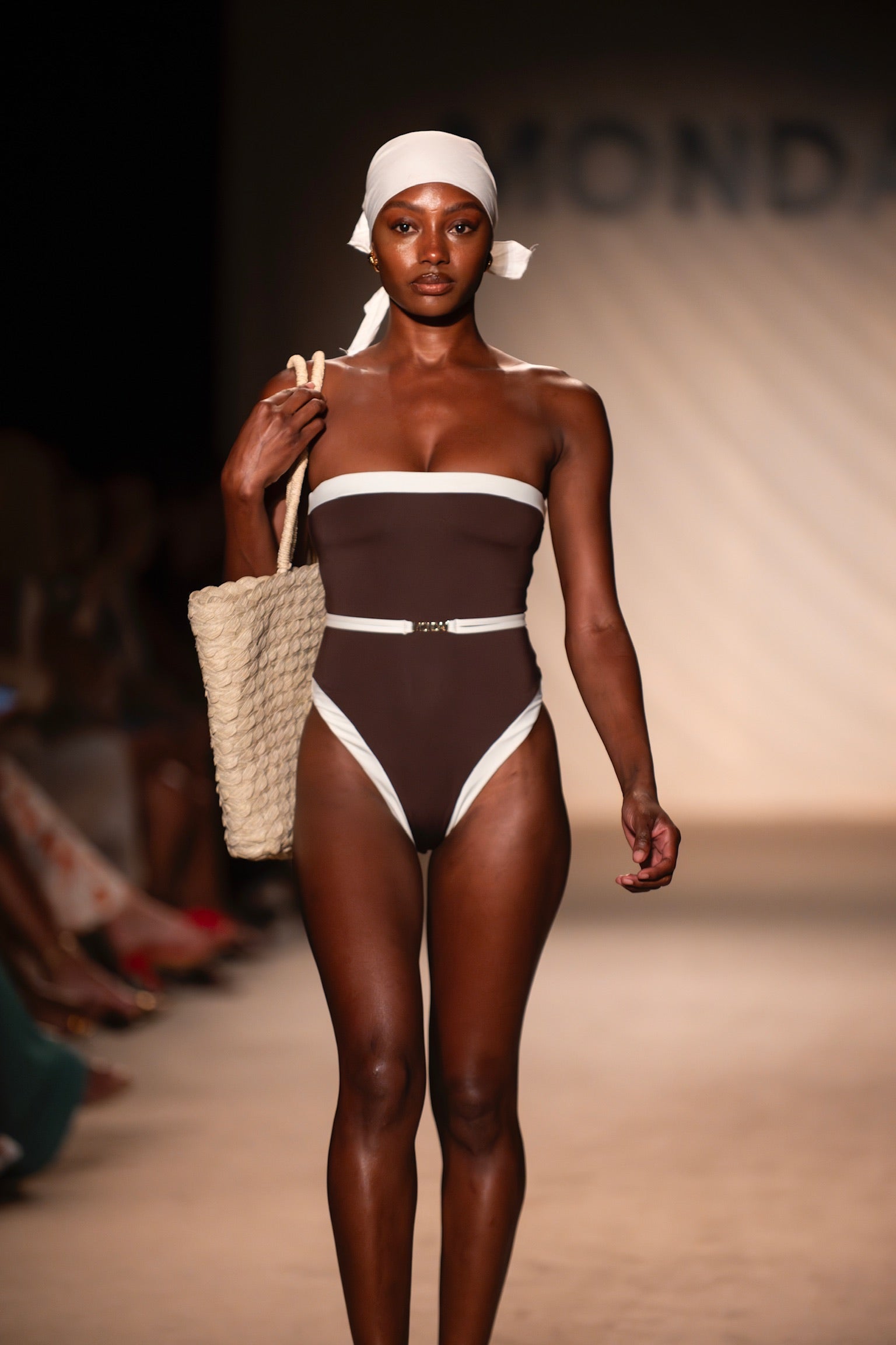 Runway model in brown strapless swimsuit with white trim, white headscarf, and woven straw tote over shoulder.