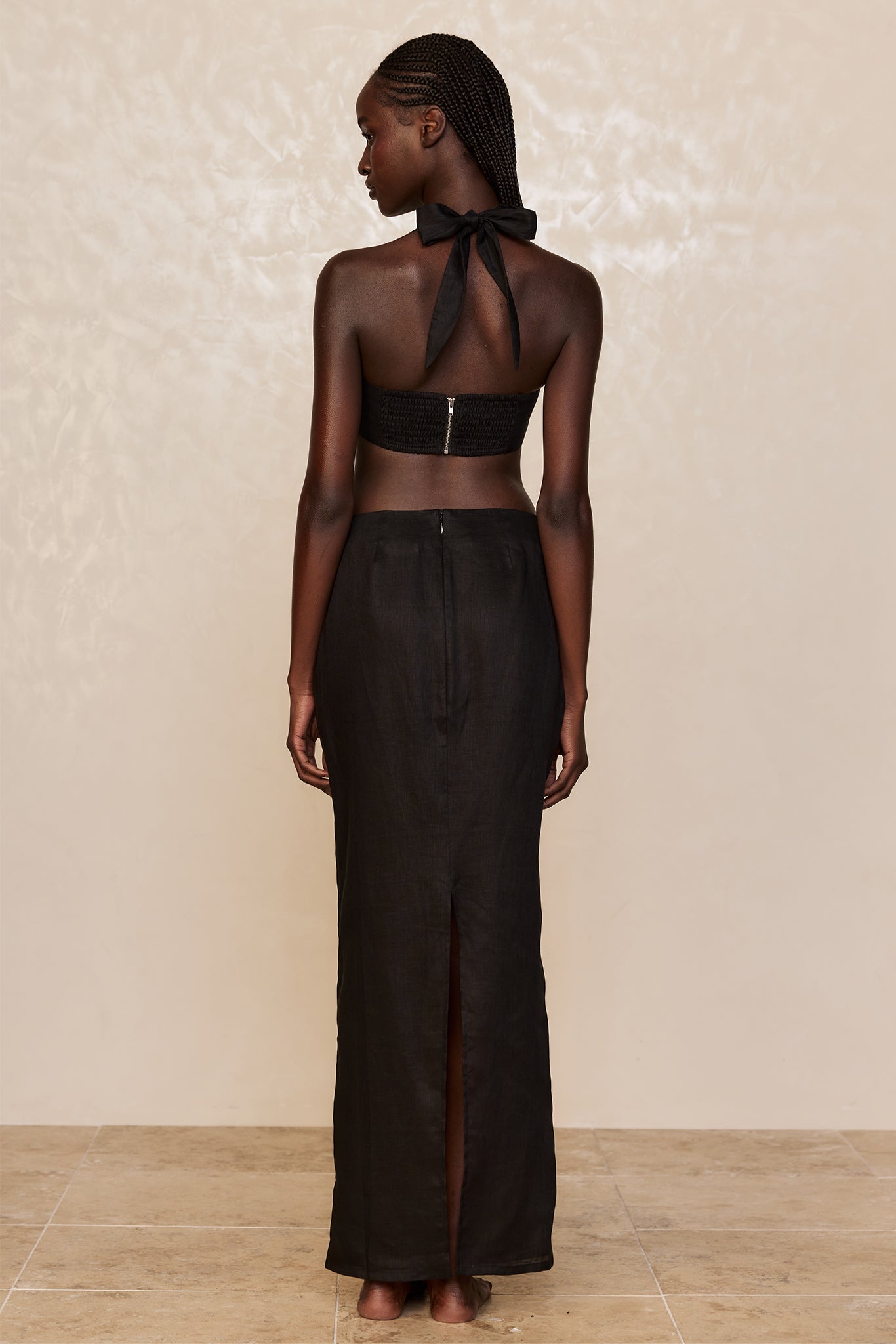 Back view of woman wearing a black bandeau top with tied neck bow and long black skirt with center back slit, bare feet