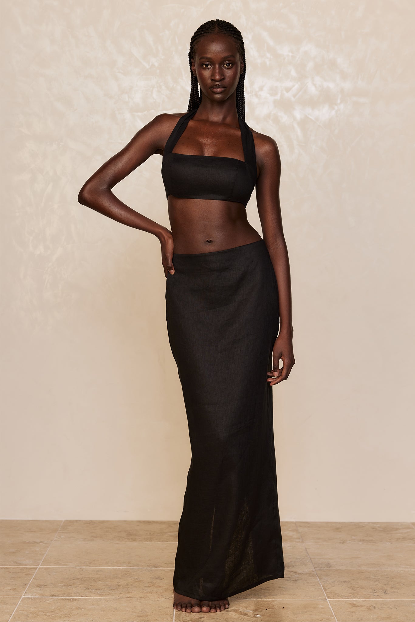 Dark-skinned model wearing a black halter crop top and long skirt, barefoot, hand on hip, neutral textured backdrop.