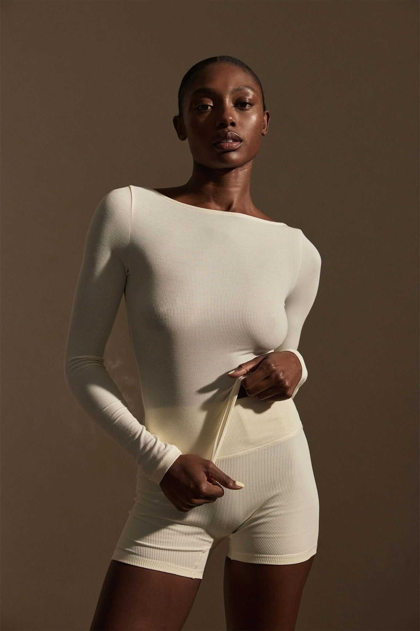 Model wearing cream ribbed long-sleeve top and matching shorts, tugging the top's hem to show fit and texture.