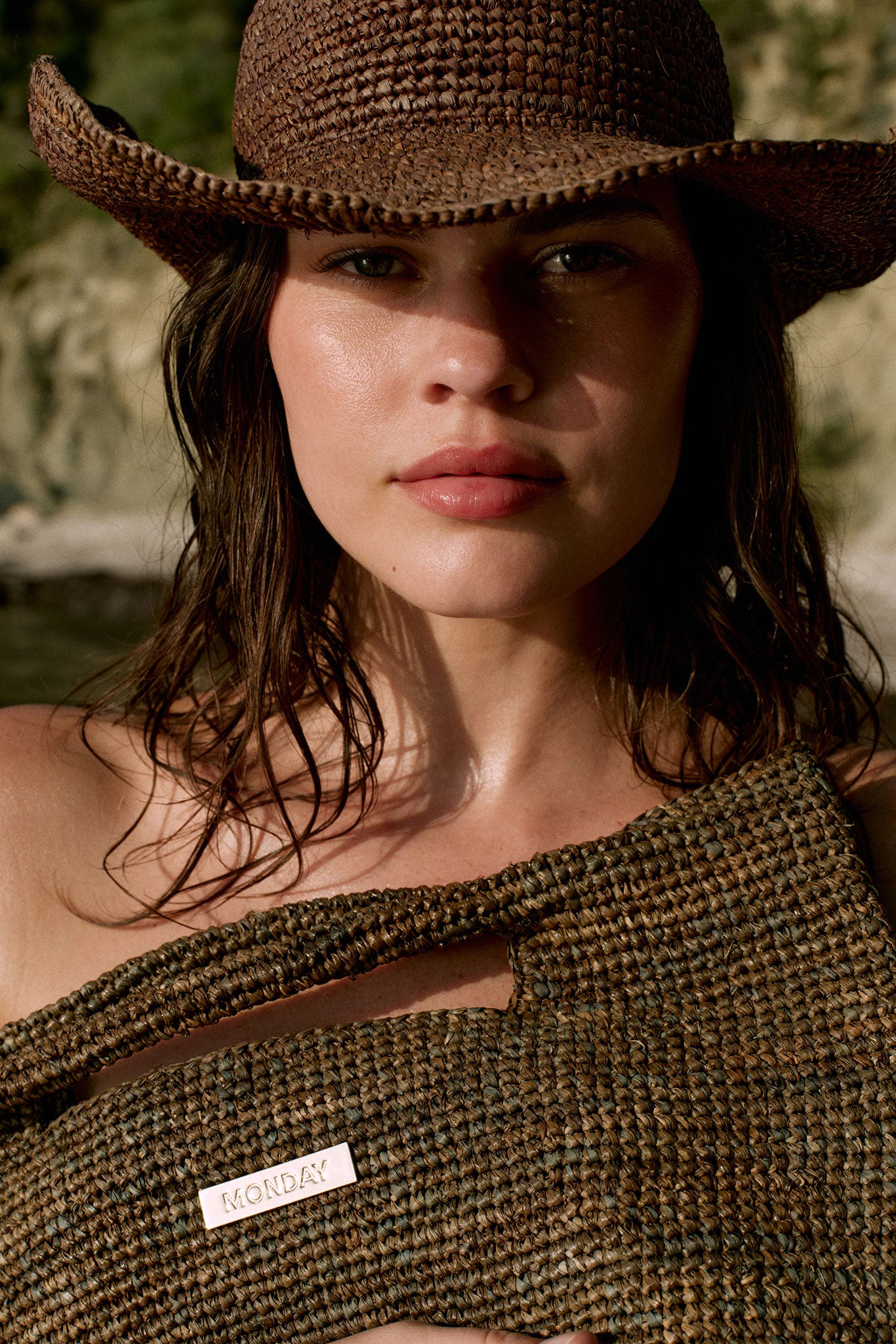 Close-up portrait of a woman in a woven sun hat holding a braided straw bag with metal tag reading MONDAY.
