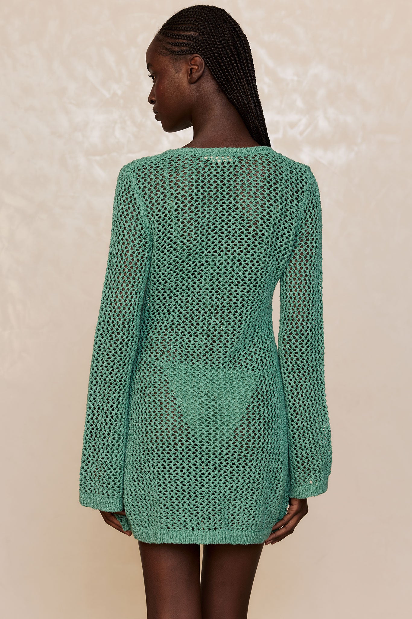 Back view of a woman with braided hair wearing a teal open-knit long-sleeve sweater dress with visible matching bikini bottom.