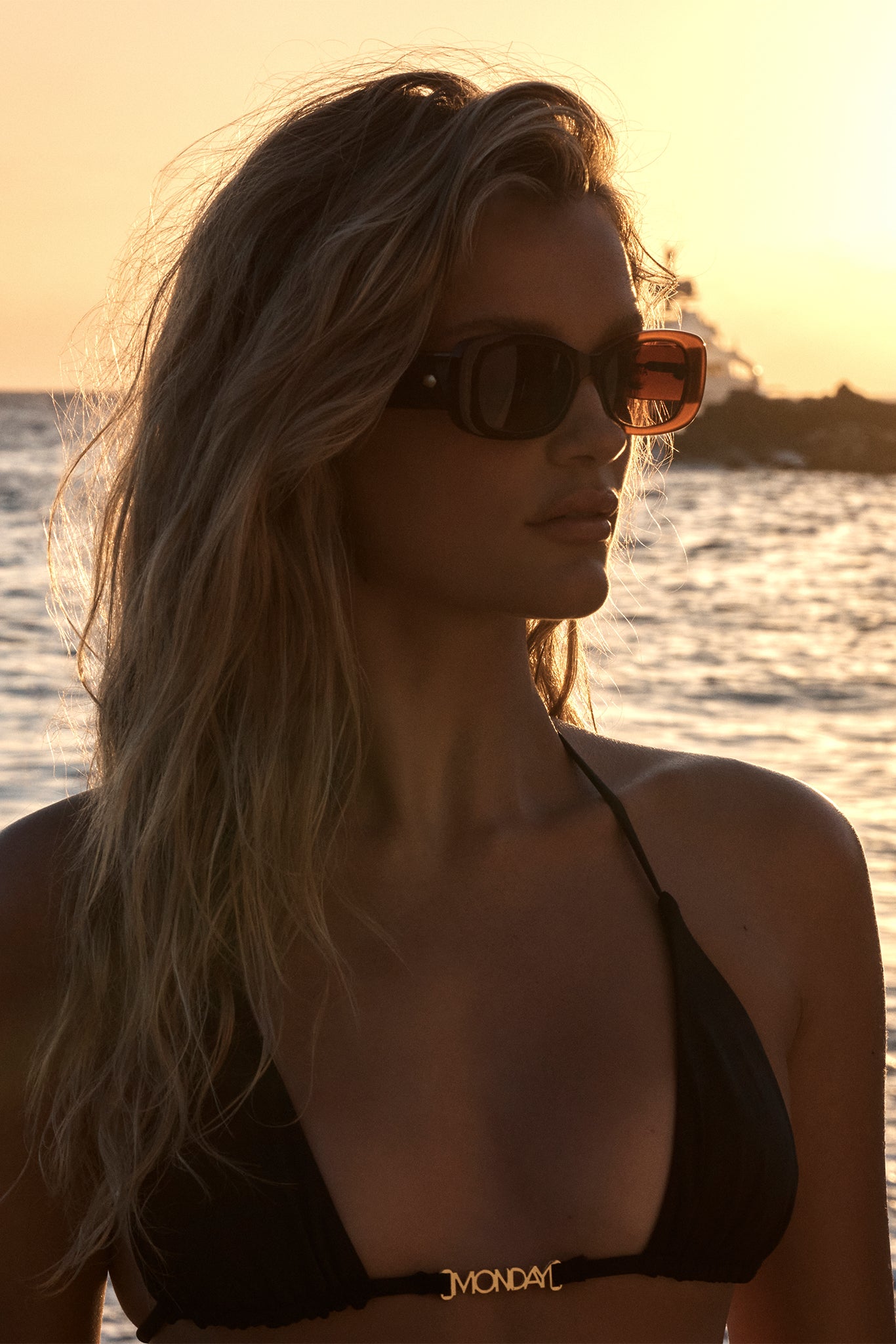 Woman wearing dark sunglasses and a black bikini top with 'JMONDAY' clasp, standing at sunset by the sea