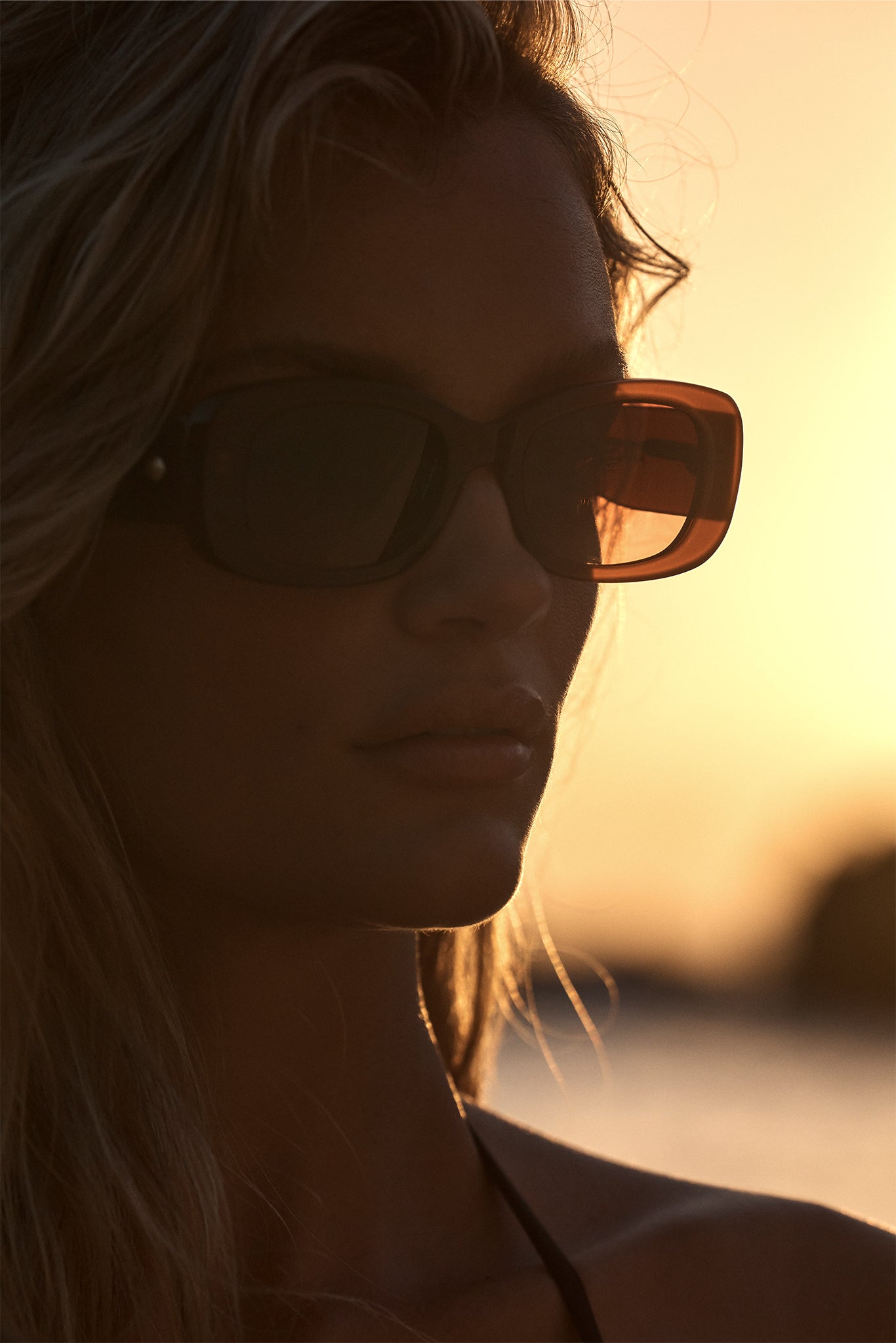 Close-up portrait of a person wearing rectangular sunglasses against a warm sunset glow