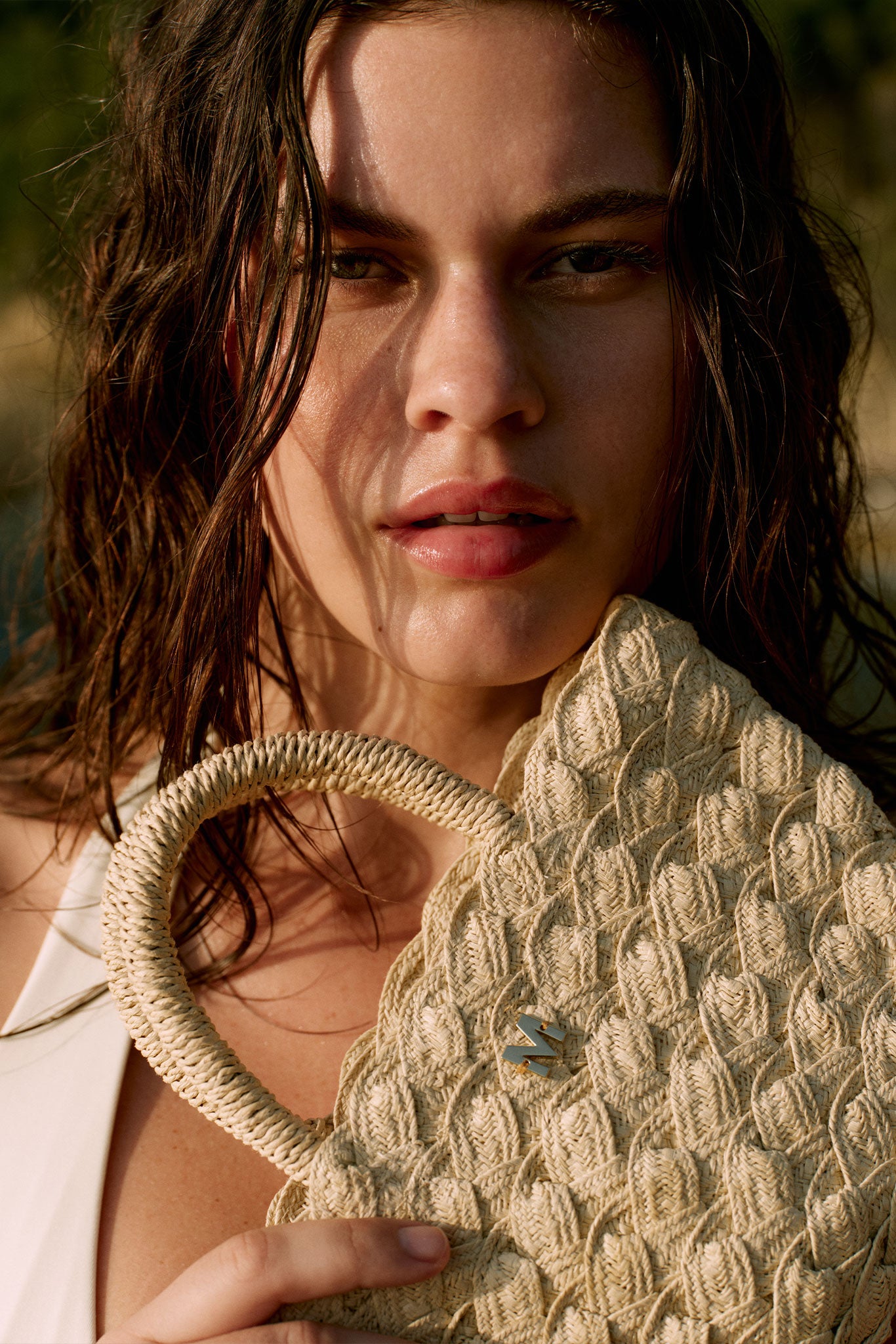 Close-up of a woman with wet hair holding a woven straw bag with braided handle, metal M logo visible on bag.
