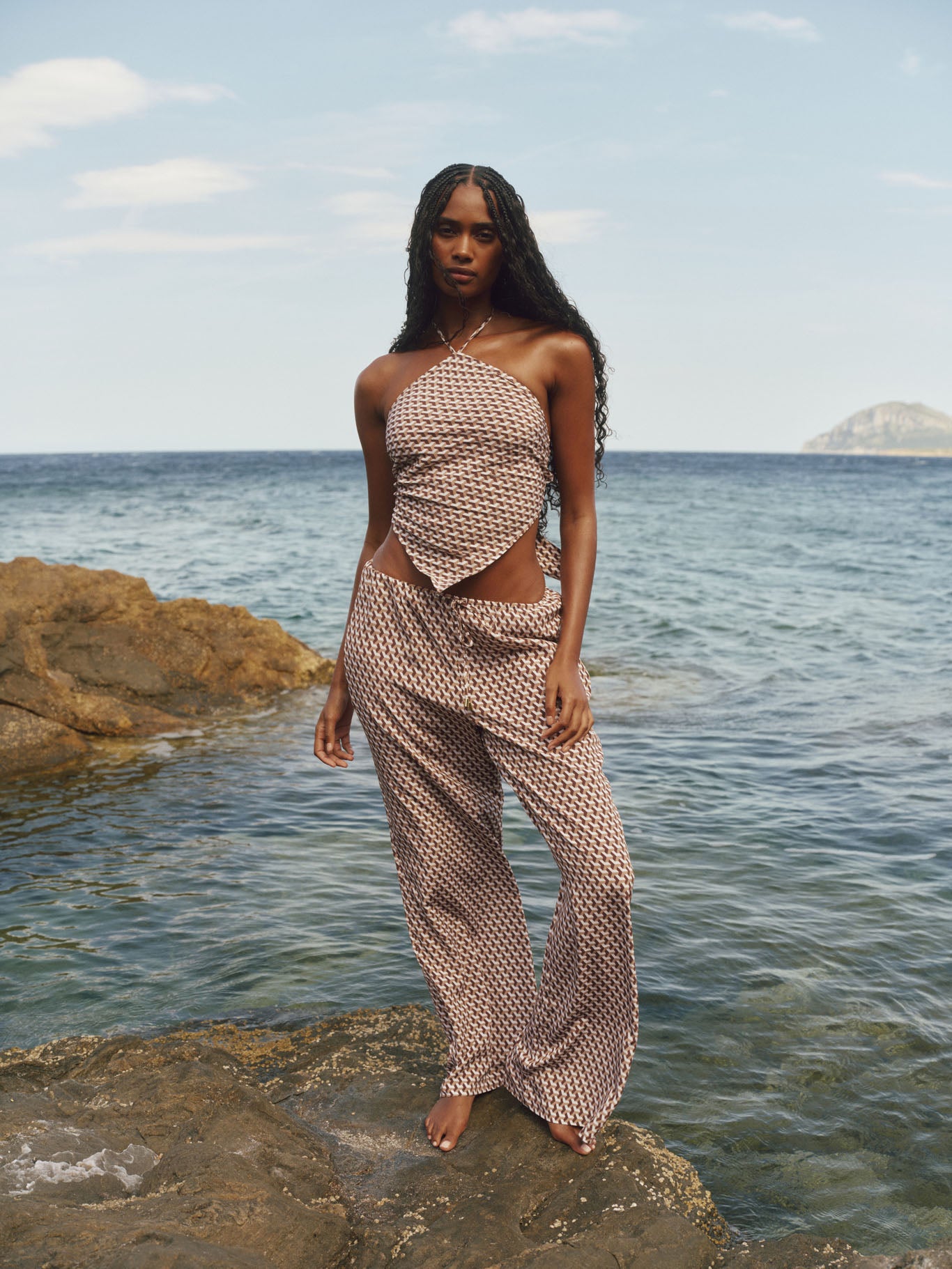 Front view of Belize Pant in Husk Geo Silk showing brown geo print, high-rise cutout waist and wide-leg fit on rocky beach