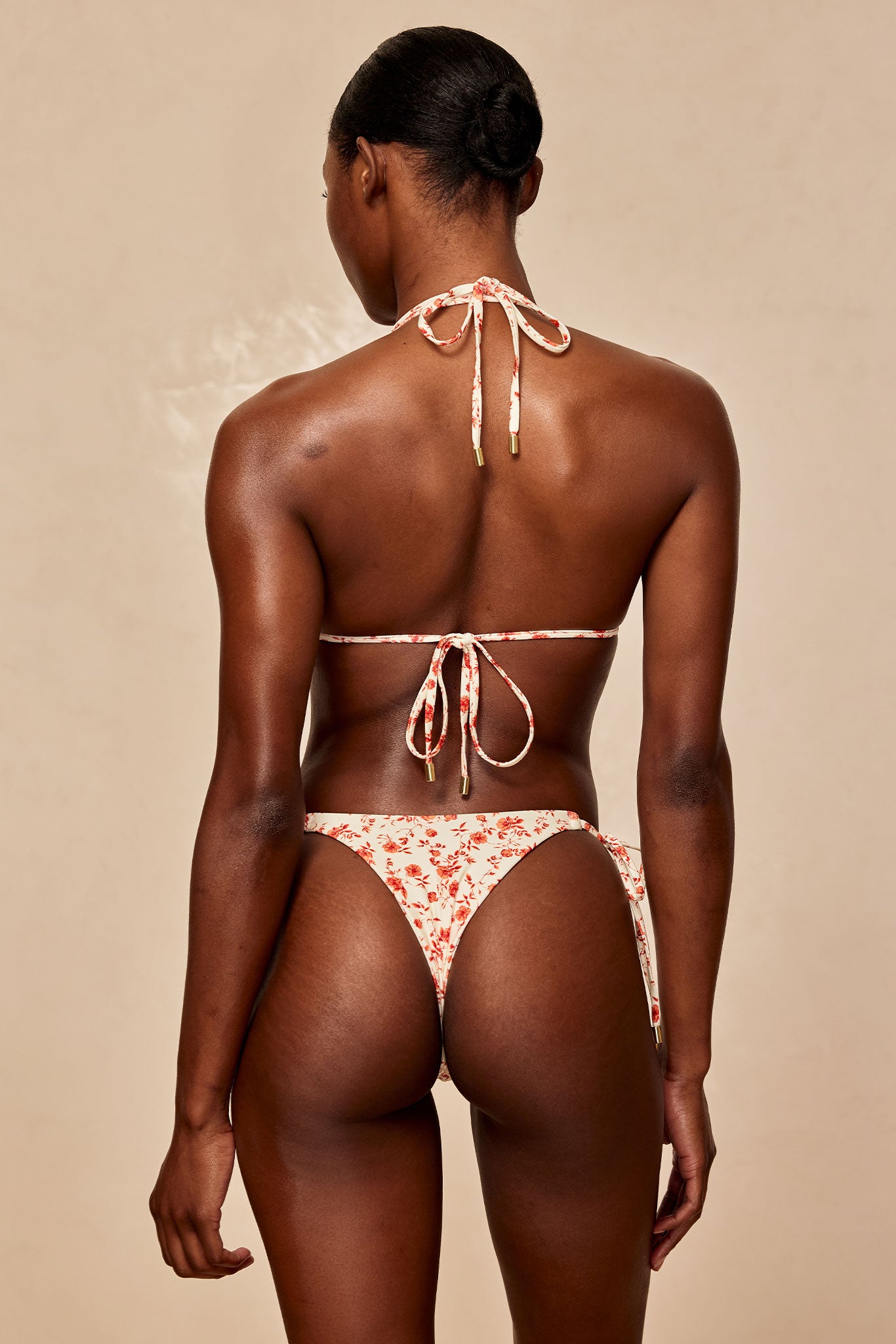 Back view of model wearing cream bikini with red floral print, halter-tie top and tie-side bottoms.