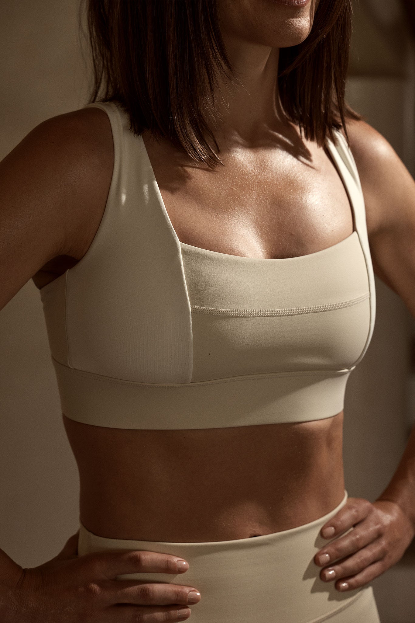 Woman's torso in beige sports bra and high-waisted leggings, hands on hips, cropped at chin