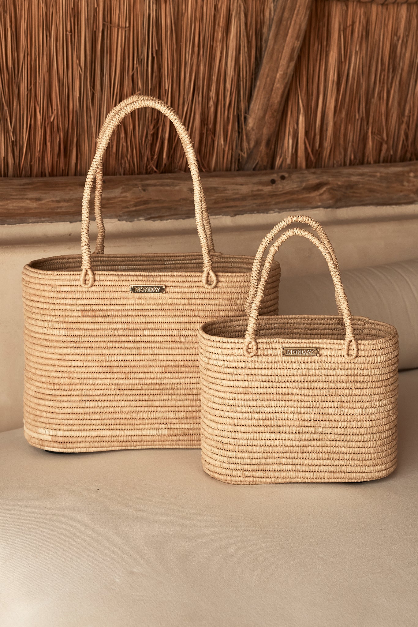 Two woven straw tote bags with loop handles and metal label MONDAY, placed on a light sofa under a thatch roof.