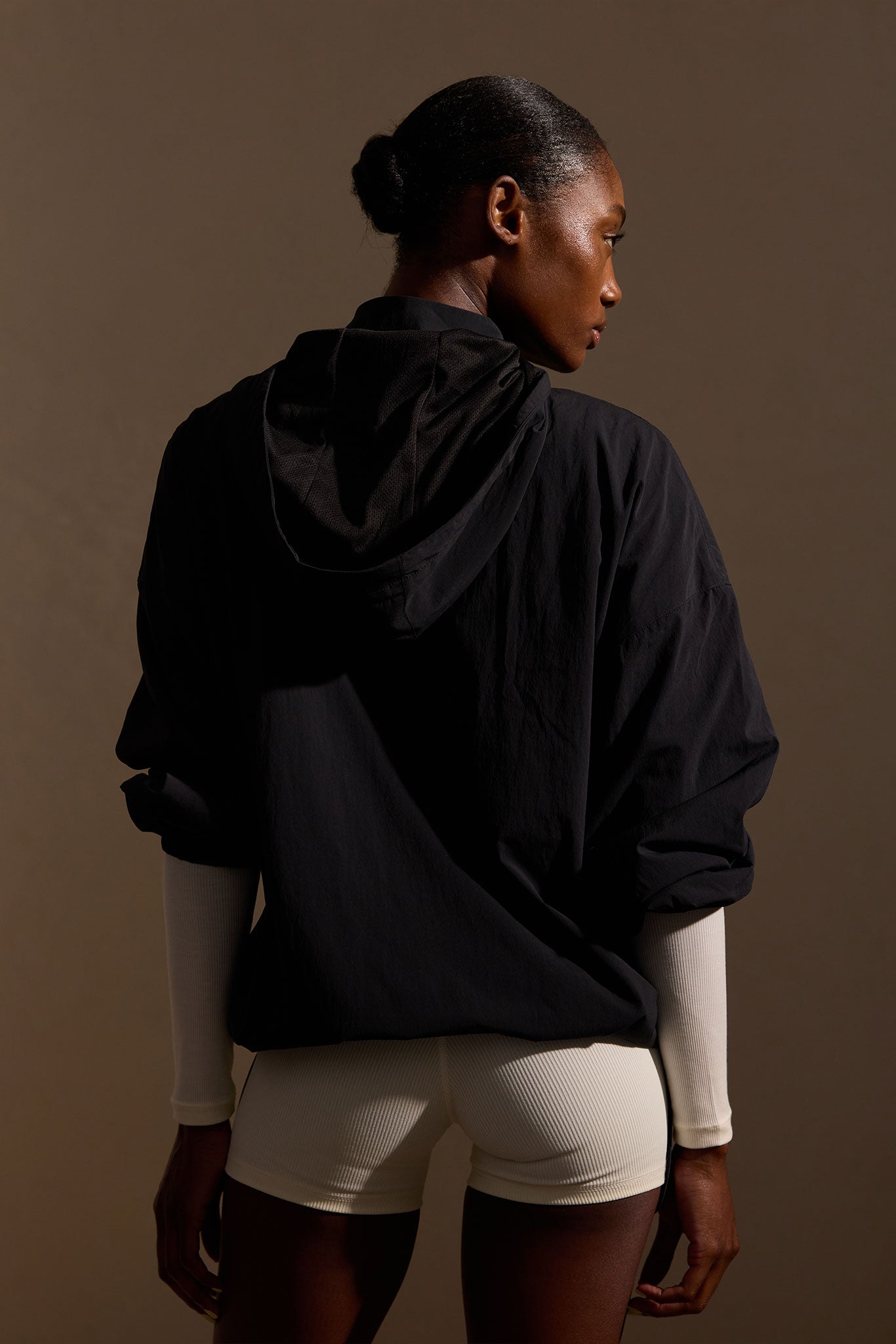 Back view of woman wearing black hooded jacket and cream ribbed shorts, hair in a bun, studio-lit, glancing over shoulder.