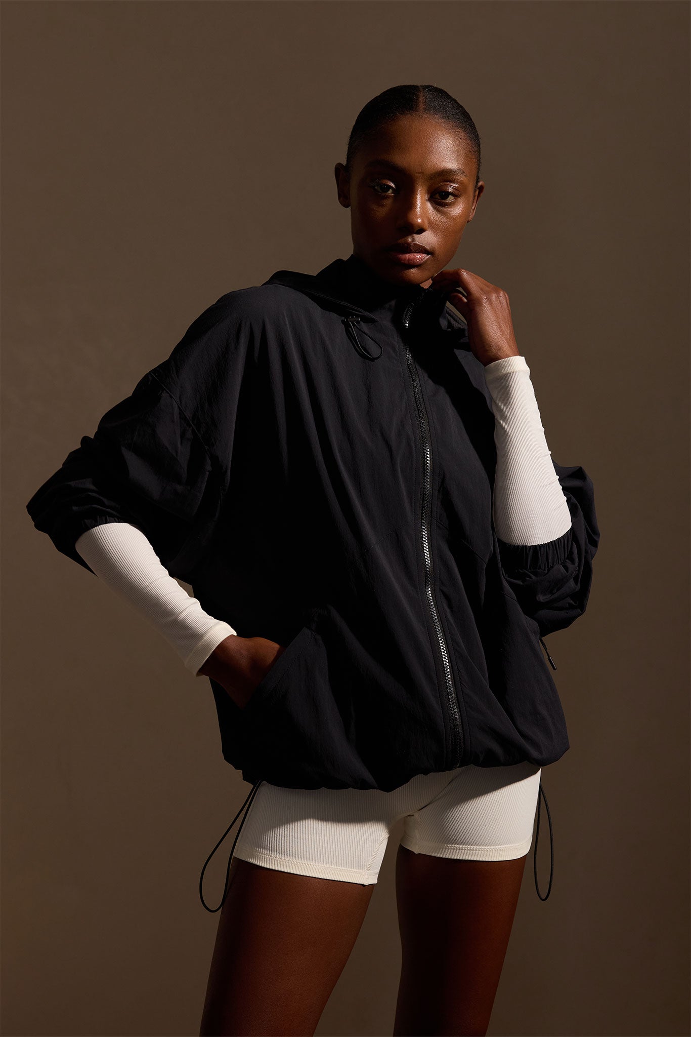 Person wearing a black zip-front hooded jacket over white long sleeves and ribbed white shorts, hand at collar, neutral brown background