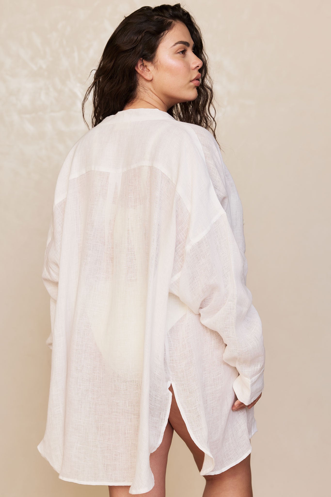 Woman wearing a sheer white long shirt cover-up, shown from the back looking over her shoulder.