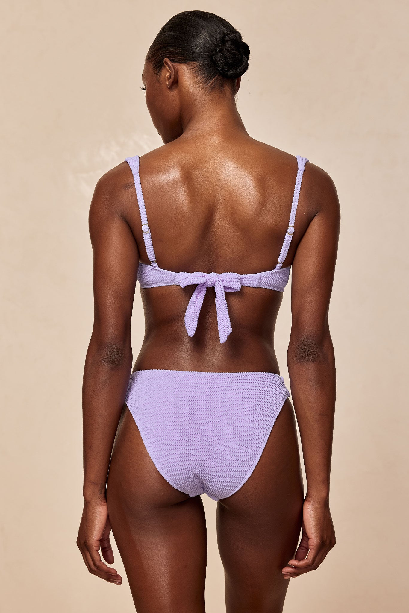 Woman seen from back wearing a lavender textured bikini with tied-back top and mid-rise bottoms; hair in a neat bun.