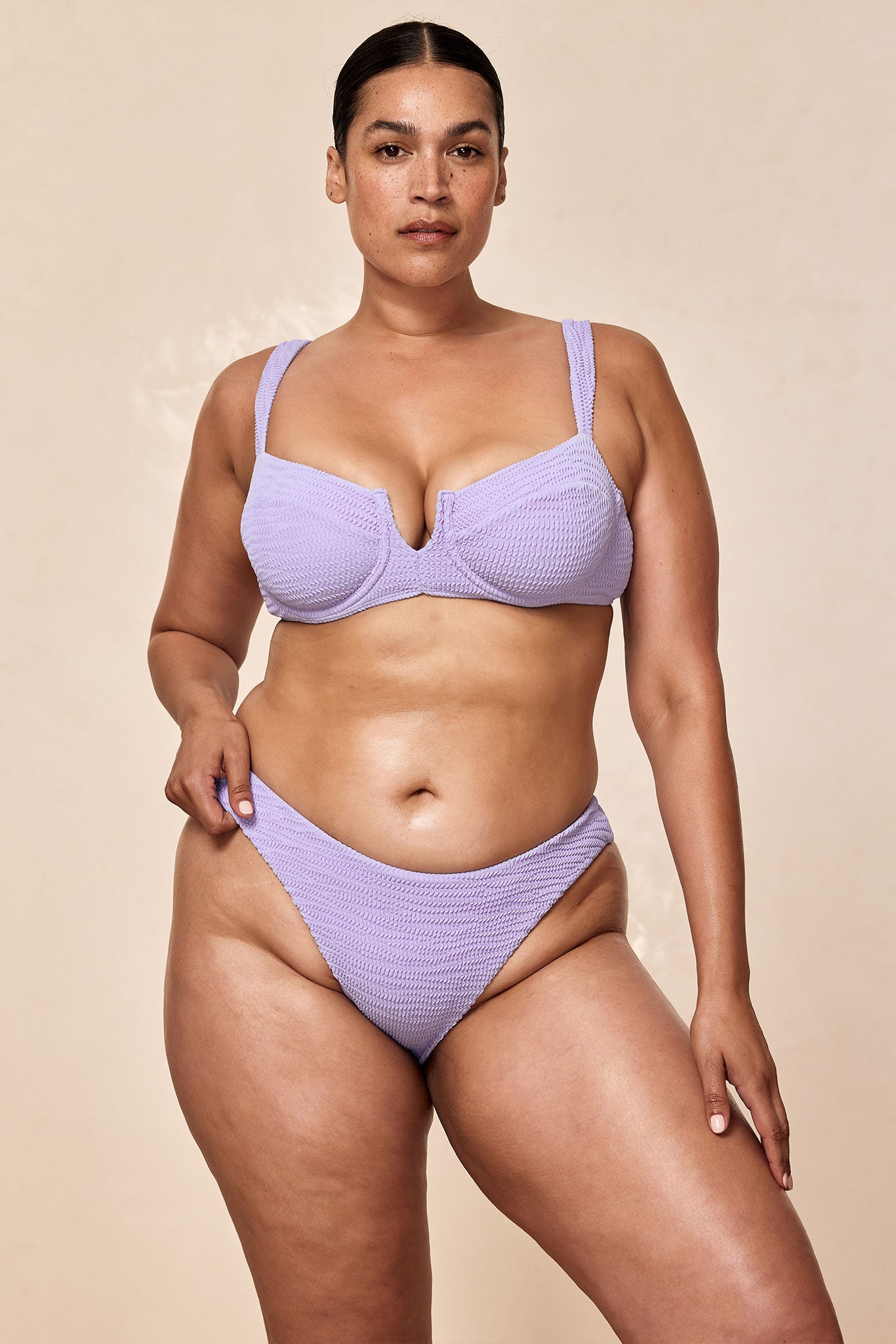 Plus-size model in lavender textured underwire bikini, standing and tugging the high-cut bottom.