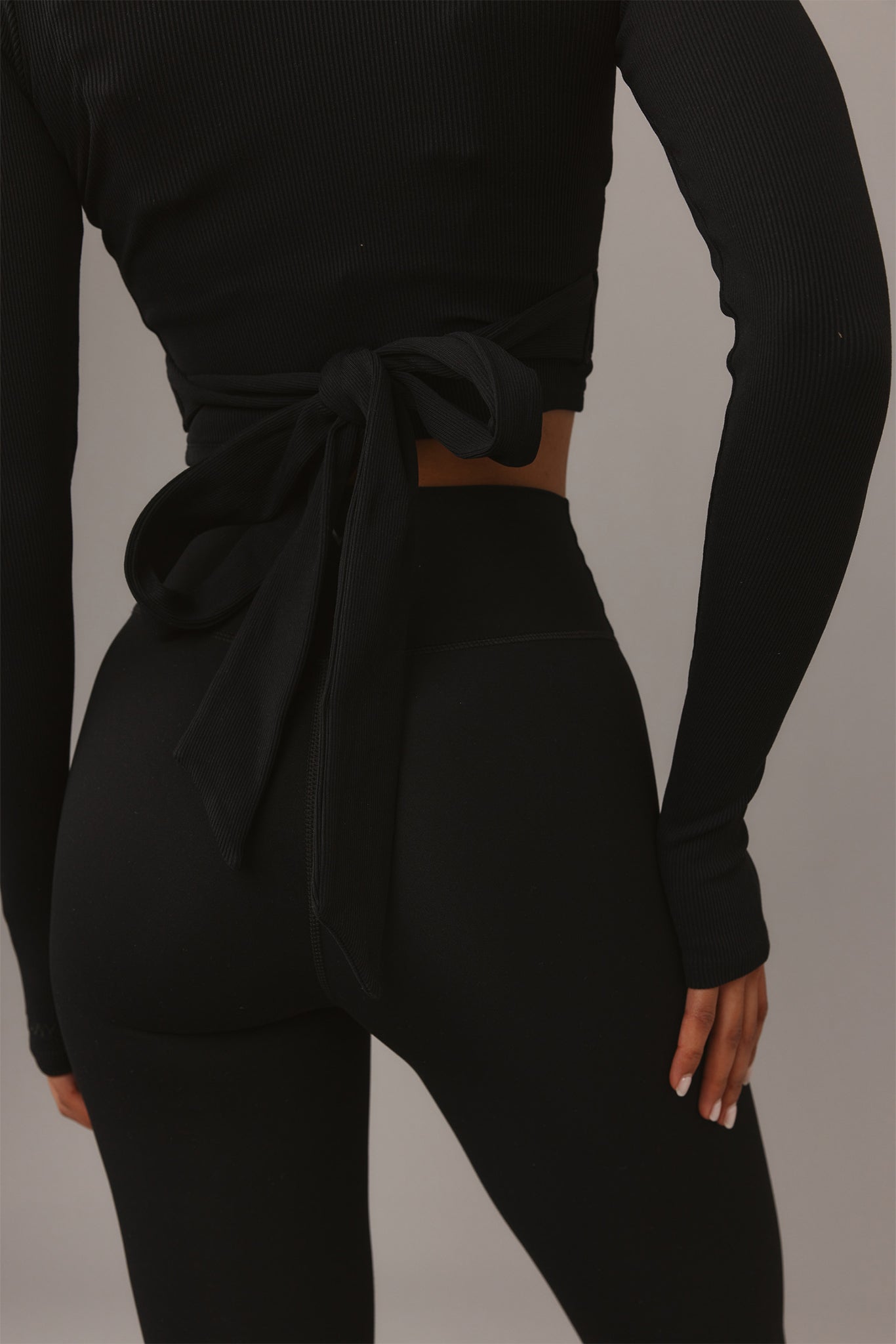 Back view of person in black ribbed wrap top tied at waist and high-waisted leggings, hand at side.