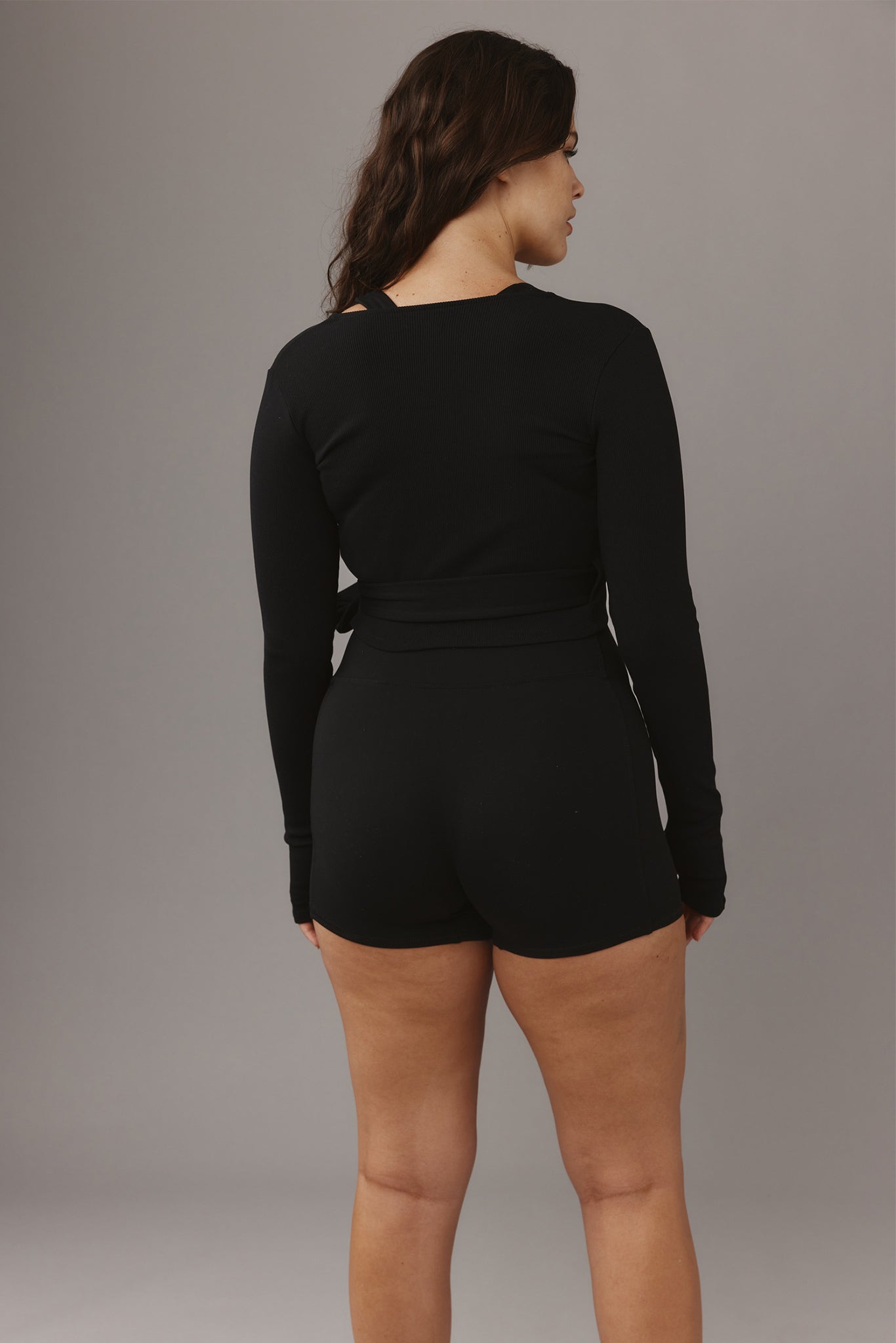 Woman seen from behind wearing a black long-sleeve wrap top and fitted high-waisted black shorts against a gray backdrop.