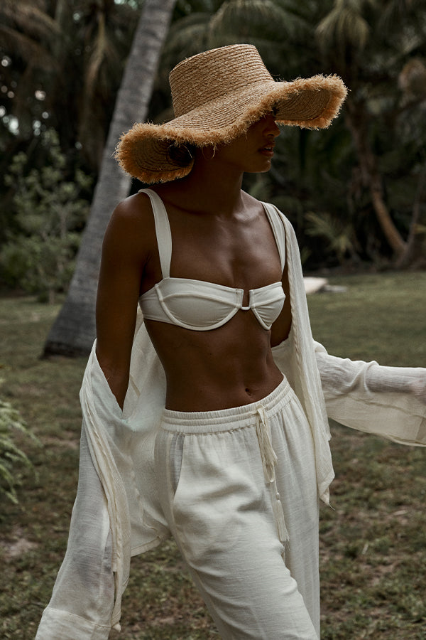 Person in straw sun hat, white bikini-style top and loose white pants standing on grassy area with palm trees