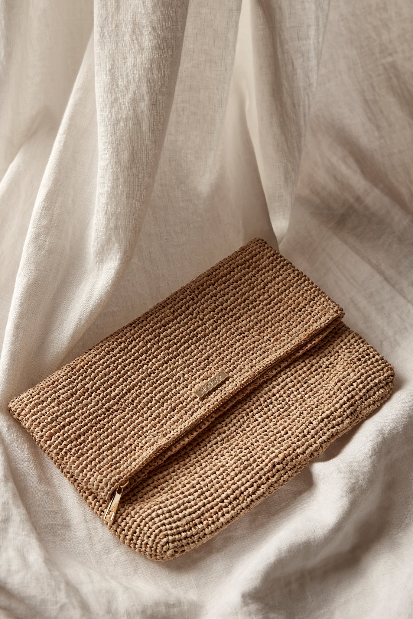 Woven tan raffia clutch with MANGO metal logo plaque, resting on draped cream linen fabric