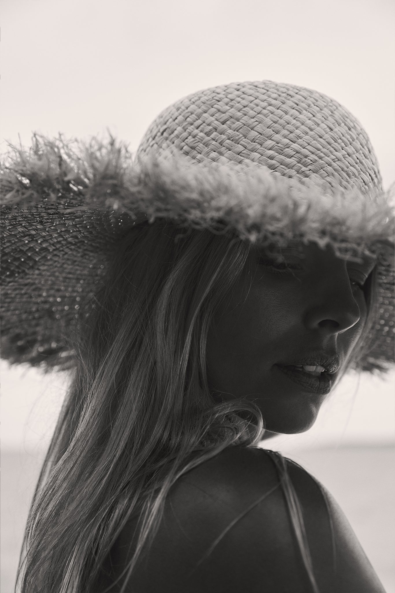 Black-and-white close-up of a woman turned over her shoulder wearing a wide-brim woven straw hat on the beach.