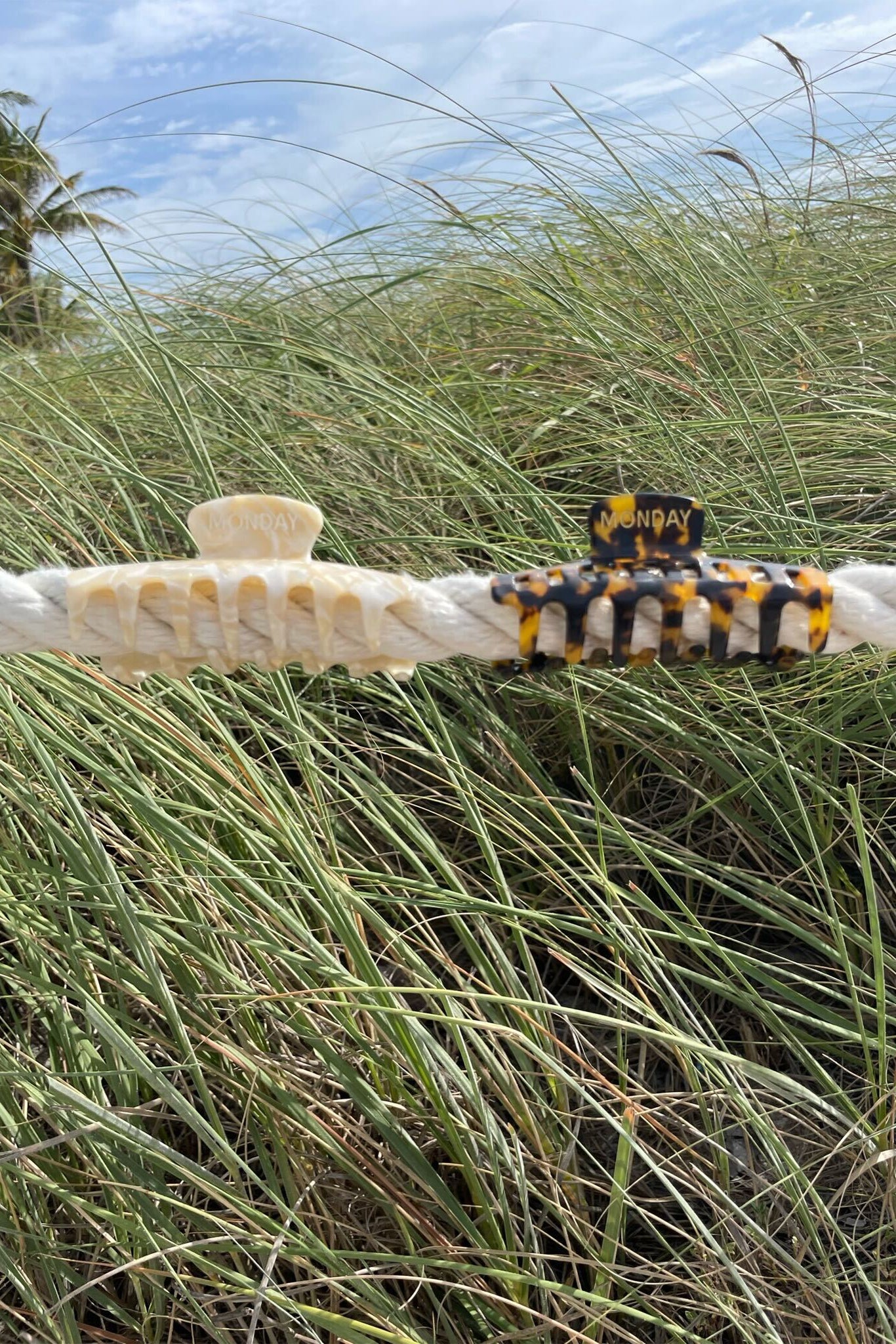 Two small hair claw clips labeled 'MONDAY' clipped to a white rope against dune grass and blue sky.