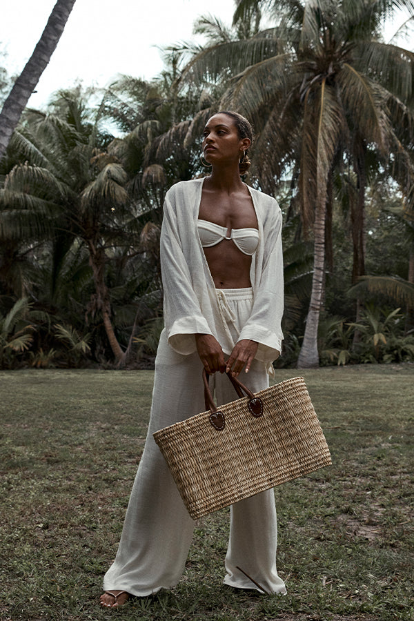 Woman in white lounge set and bikini top holding woven straw tote in a palm-lined garden