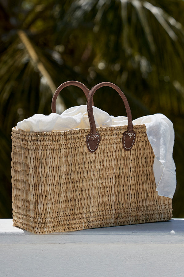 Rectangular woven straw tote bag with brown leather handles and a white linen scarf draped inside