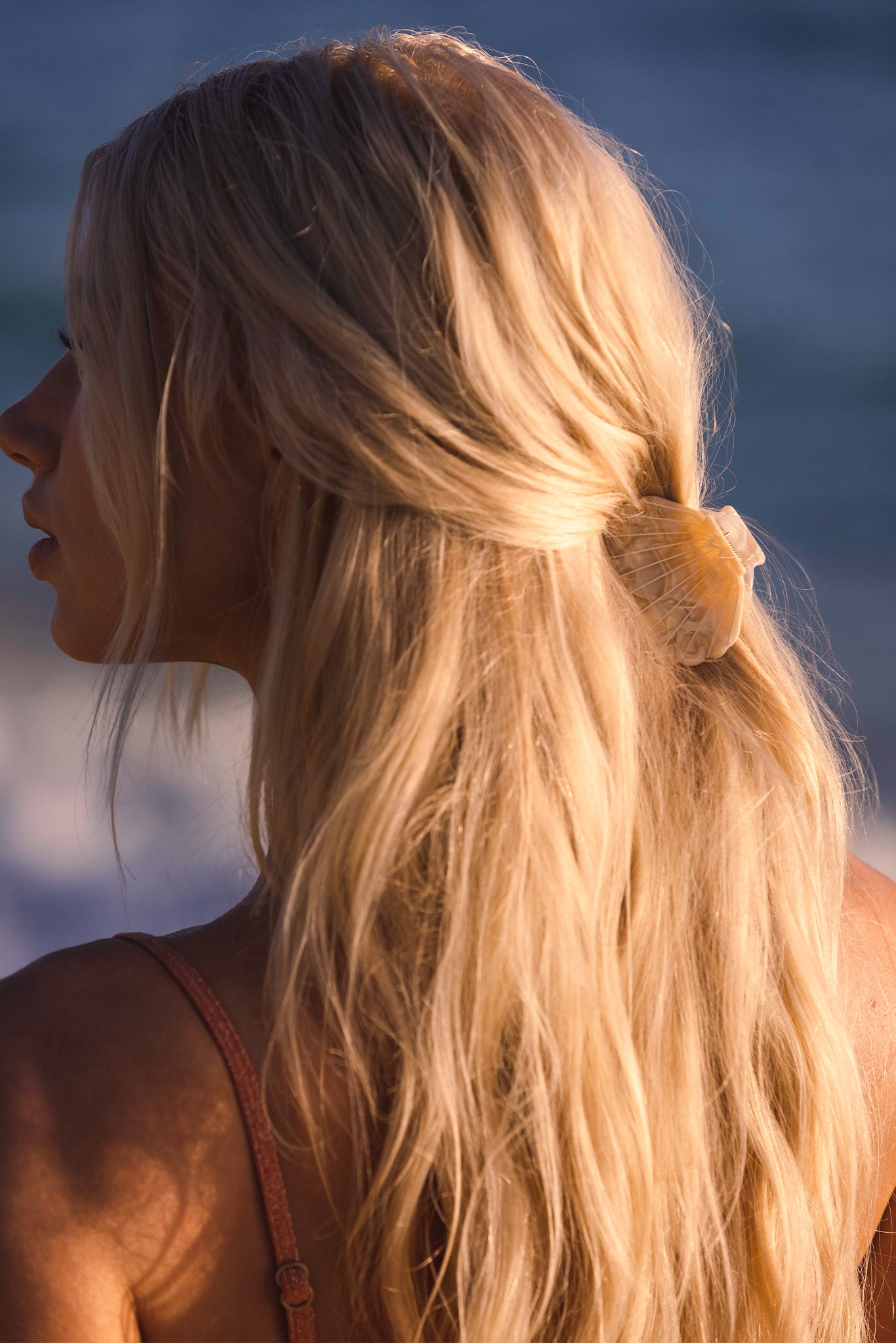 Back view of a woman with long sunlit blonde hair secured with a shell-shaped hair clip at the beach.
