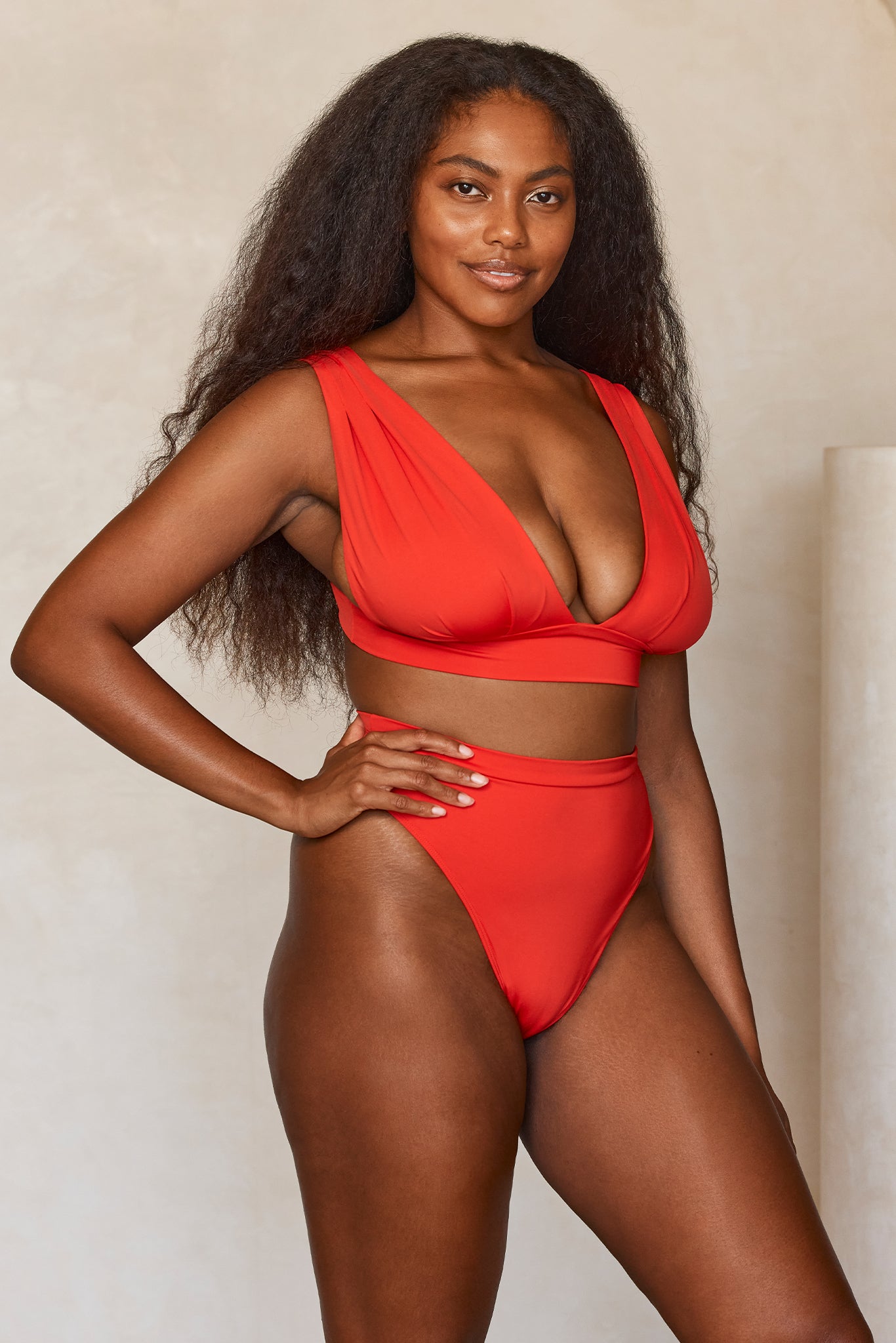 Woman with long natural hair wearing a high‑waist red bikini, hand on hip, smiling against a neutral background.