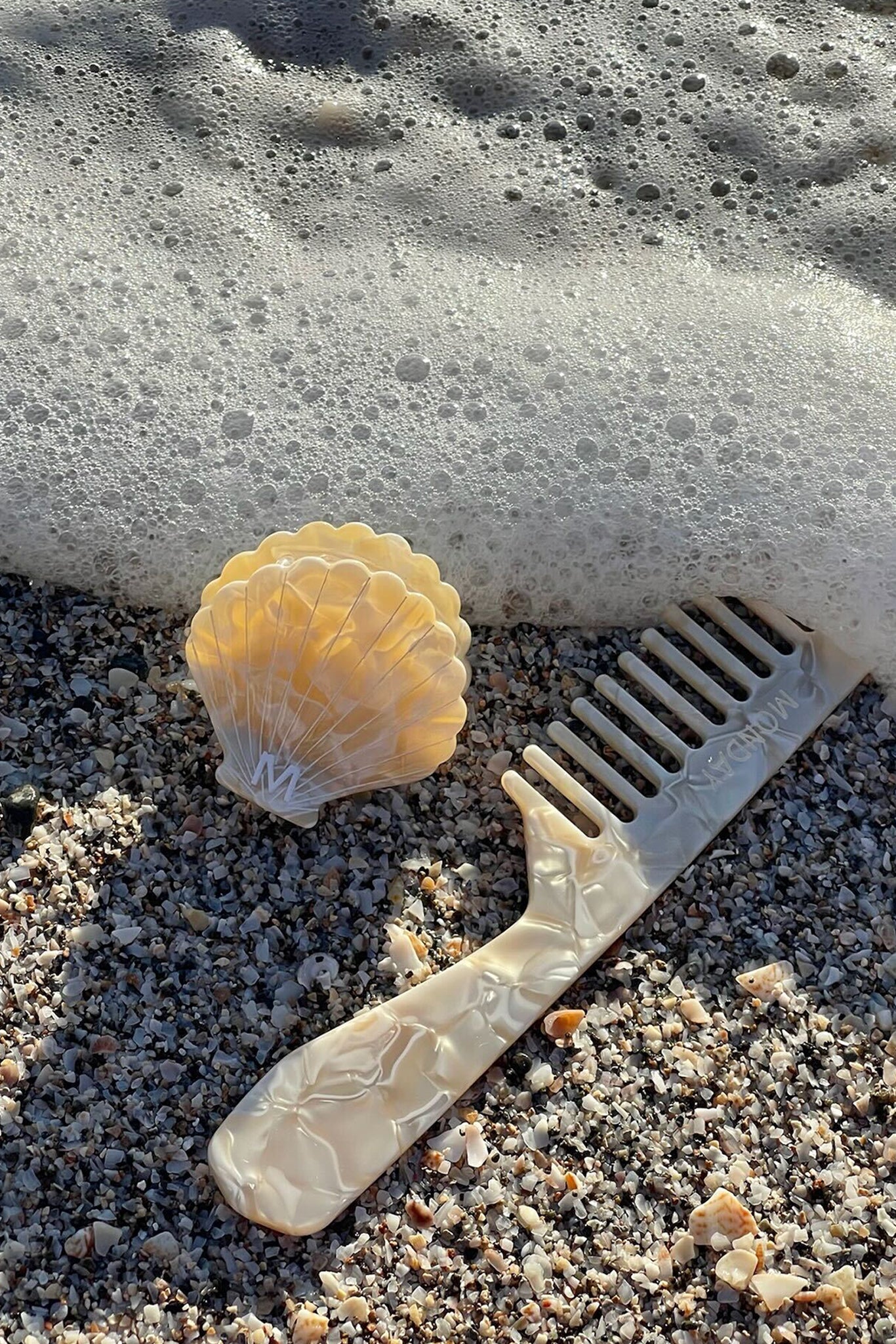 Pearlescent shell-shaped hair clip and matching wide-tooth comb on pebbly beach with foamy surf