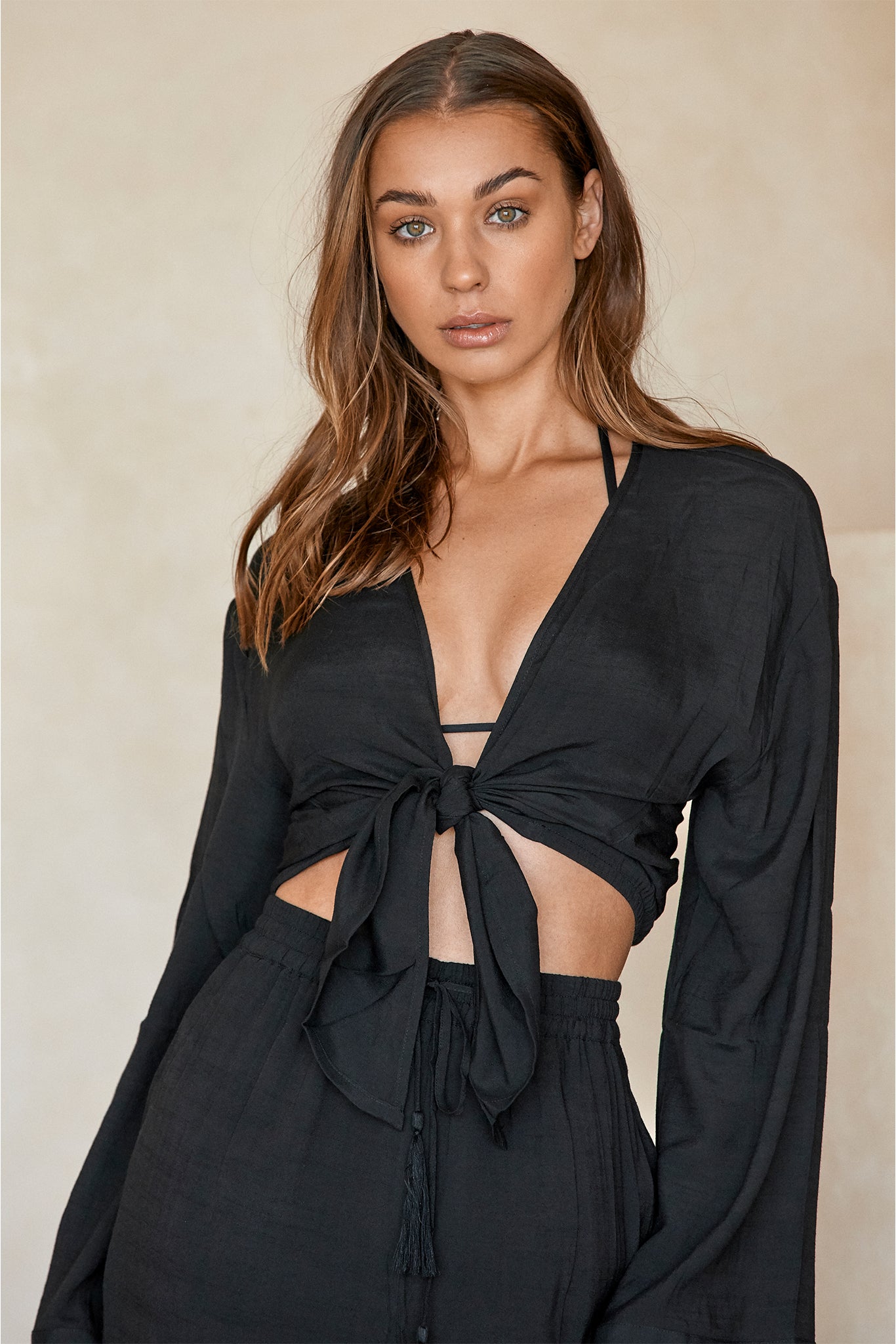 Woman wearing black tie-front crop top and matching high-waist pants, facing camera against a neutral beige background.